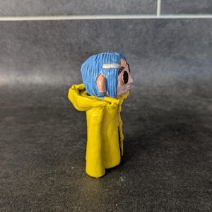 Puede incluir: Figura hecha a mano de un personaje con cabello azul, vistiendo un impermeable amarillo. La figura tiene una cara detallada con ojos negros y una diadema blanca. La figura est&aacute; sobre una superficie oscura.