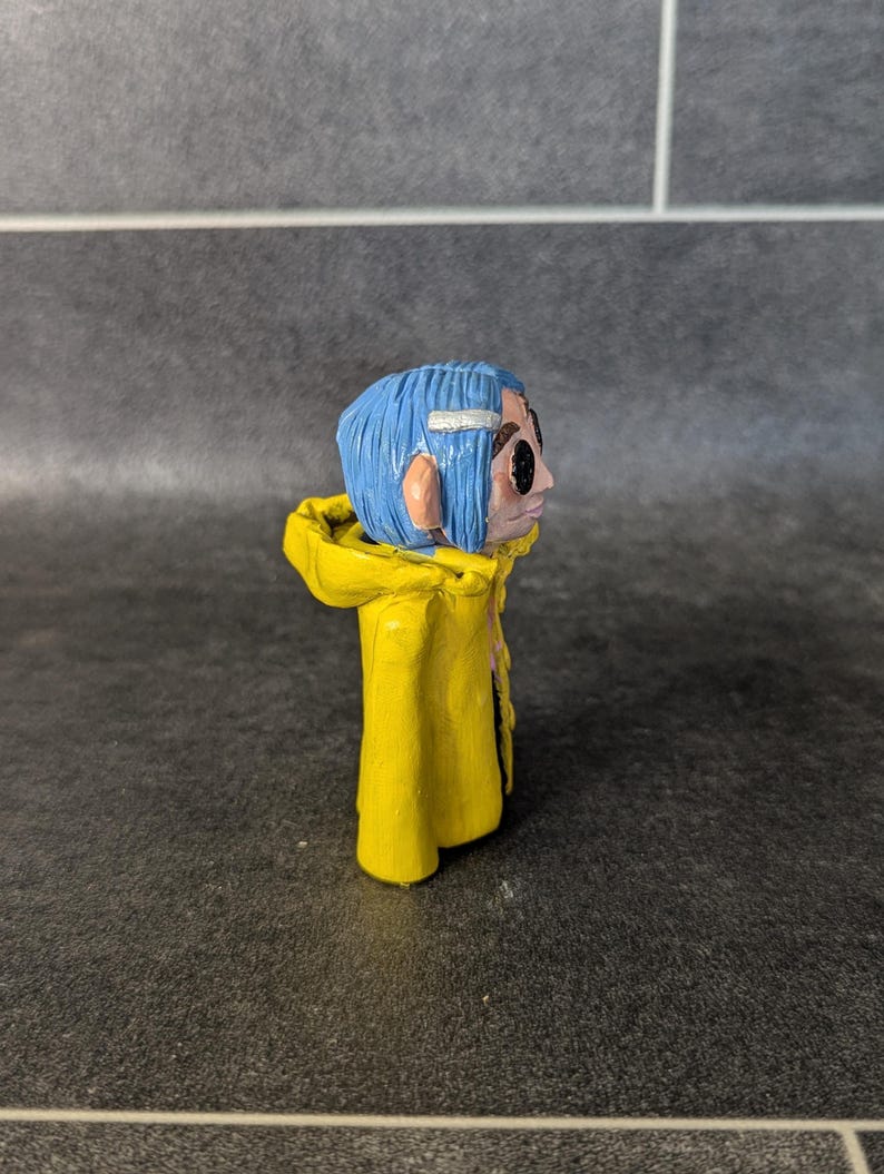 Puede incluir: Figura hecha a mano de un personaje con cabello azul, vistiendo un impermeable amarillo. La figura tiene una cara detallada con ojos negros y una diadema blanca. La figura est&aacute; sobre una superficie oscura.