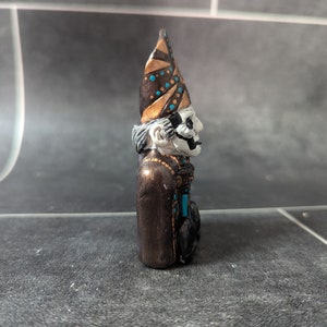Puede incluir: Una peque&ntilde;a figurita pintada a mano de una figura esquel&eacute;tica que lleva un sombrero puntiagudo con detalles dorados y marrones. La figura lleva una t&uacute;nica marr&oacute;n con detalles turquesa.