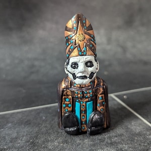 Könnte beinhalten: Eine kleine, handbemalte Figur eines Wesens, das eine goldene und schwarze Mitra mit türkisfarbenen Akzenten trägt. Die Figur hat ein weißes Gesicht mit schwarzen Augen und einem schwarzen Mund.