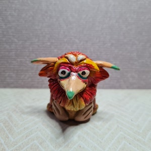 May include: A hand-painted figurine of a fantastical creature with a long beak and large eyes. The sculpture features a vibrant colour palette of red, orange, yellow, and green. The creature has horns and a fluffy mane.