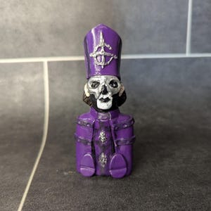 May include: A small purple figurine of a skeleton wearing a purple robe and a tall purple hat with a silver symbol. The skeleton has a silver skull and a silver cross on its chest.