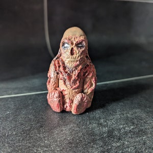 Puede incluir: Una pequeña figura detallada de una criatura con un cuerpo carnoso, rojo y marrón. La criatura tiene una cara humanoide con ojos blancos y una boca abierta de par en par.