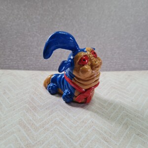 Op de afbeelding: Een blauwe en bruine cartoon-wormfiguur met rode ogen en een rode sjaal. De worm heeft een blauw, puntig oor op zijn kop.