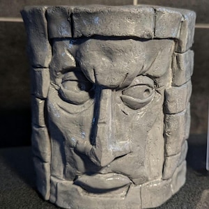 May include: A gray stone sculpture of a face with a furrowed brow and a stern expression. The sculpture is made to look like a stone block with a face carved into it.