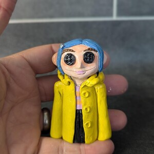 Puede incluir: Figura hecha a mano de un personaje con cabello azul, ojos de bot&oacute;n y un abrigo amarillo. La figura lleva una camisa a rayas moradas y blancas y pantalones negros. La figura se sostiene en una mano.