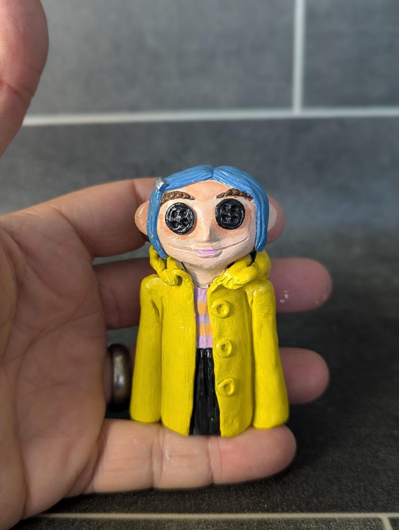 Puede incluir: Figura hecha a mano de un personaje con cabello azul, ojos de bot&oacute;n y un abrigo amarillo. La figura lleva una camisa a rayas moradas y blancas y pantalones negros. La figura se sostiene en una mano.