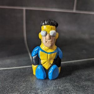 Puede incluir: Una figurita pintada a mano de un superhéroe que lleva un traje amarillo y azul con un cinturón negro. La figurita tiene el pelo negro y lleva gafas blancas.