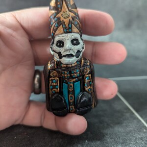 Puede incluir: Una peque&ntilde;a figurita pintada a mano de una figura que lleva una t&uacute;nica negra y dorada con una mitra azul y dorada. La figura tiene una cara blanca con ojos negros y una boca negra.