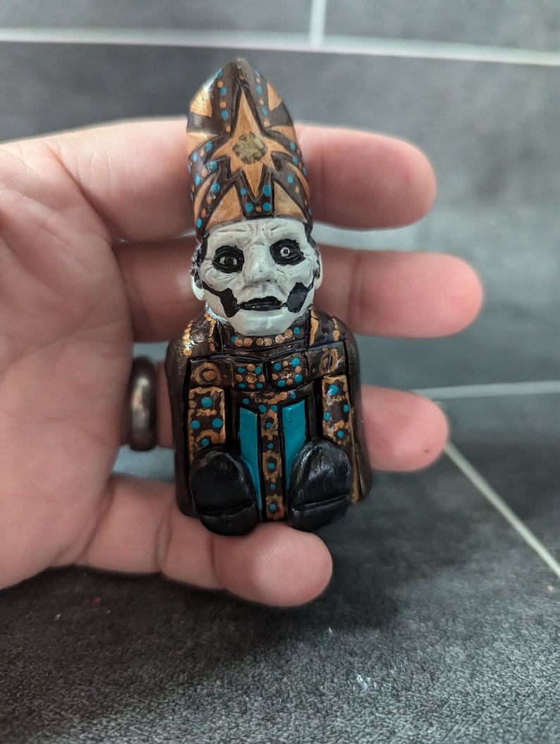 Puede incluir: Una peque&ntilde;a figurita pintada a mano de una figura que lleva una t&uacute;nica negra y dorada con una mitra azul y dorada. La figura tiene una cara blanca con ojos negros y una boca negra.