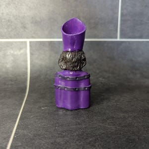 May include: Purple plastic figurine of a person wearing a purple hat and robe with black accents. The figurine is standing on a gray tiled surface.