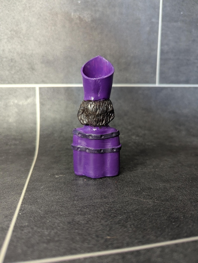 May include: Purple plastic figurine of a person wearing a purple hat and robe with black accents. The figurine is standing on a gray tiled surface.