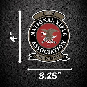 May include: A circular National Rifle Association Life Member patch featuring a central red field with a brown eagle and crossed rifles, surrounded by black text on a tan border. The patch measures 10.16 cm in height and 8.26 cm in width.