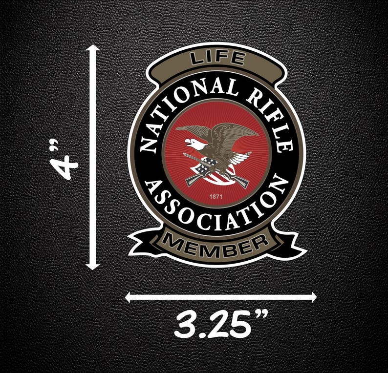 May include: A circular National Rifle Association Life Member patch featuring a central red field with a brown eagle and crossed rifles, surrounded by black text on a tan border. The patch measures 10.16 cm in height and 8.26 cm in width.