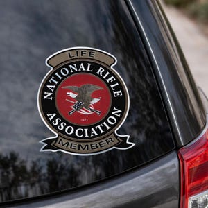 May include: A circular National Rifle Association Life Member decal featuring a red centre with a bald eagle and crossed rifles graphic. The black border displays the text National Rifle Association, with tan banners reading Life and Member.