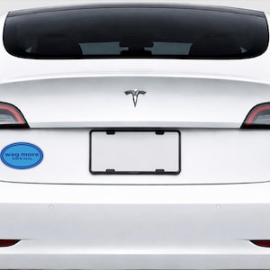 May include: A white Tesla car with a blue oval bumper sticker that reads "wag more, bark less." The car has a black license plate frame and a Tesla logo.