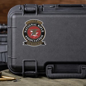 May include: A black rectangular hard-shell carrying case featuring a circular National Rifle Association Life Member sticker. The sticker displays a central eagle emblem, crossed rifles, and the text National Rifle Association Life Member.