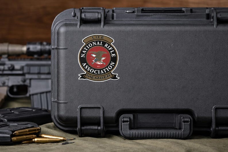 May include: A black rectangular hard-shell carrying case featuring a circular National Rifle Association Life Member sticker. The sticker displays a central eagle emblem, crossed rifles, and the text National Rifle Association Life Member.