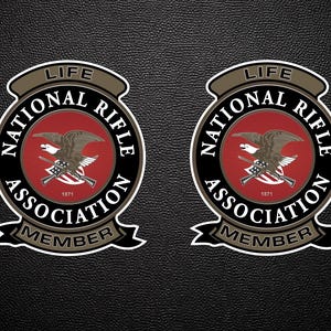 May include: Two circular National Rifle Association Life Member emblems featuring a central red field with a brown eagle and crossed rifles. The text reads LIFE, NATIONAL RIFLE ASSOCIATION, and MEMBER, with the year 1871 printed in white.