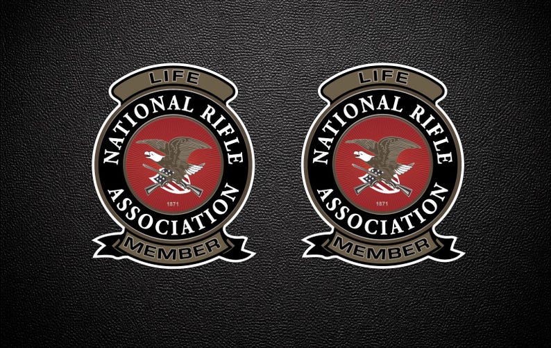 May include: Two circular National Rifle Association Life Member emblems featuring a central red field with a brown eagle and crossed rifles. The text reads LIFE, NATIONAL RIFLE ASSOCIATION, and MEMBER, with the year 1871 printed in white.