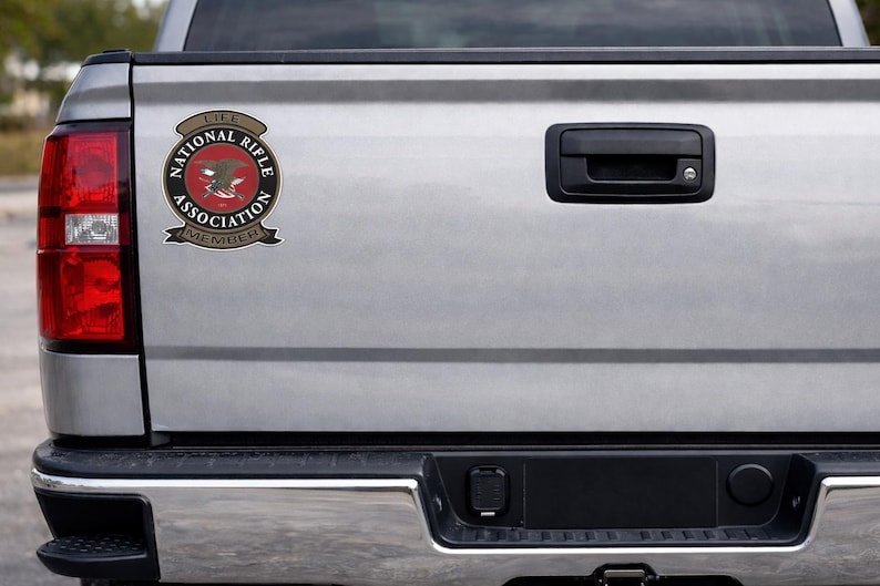 May include: A circular National Rifle Association Life Member decal featuring a red centre with a black eagle emblem, surrounded by a black border with white text, and tan banners above and below reading Life and Member respectively.