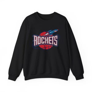 May include: Black crew neck sweatshirt featuring the word "Rockets" in white and blue lettering over a red basketball. A blue and red rocket with a shark graphic is above the word "Rockets".