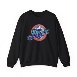 Puede incluir: Sudadera negra de cuello redondo con el logo de Utah Jazz. El logo es un diseño circular con la palabra "JAZZ" en azul y blanco, una pelota de baloncesto y la palabra "UTAH" encima de una cordillera.