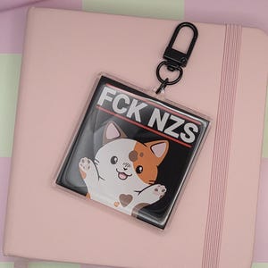 May include: A clear acrylic keychain with a black border and a black background with the text "FCK NZS" and a cartoon cat. The keychain is attached to a black metal clasp. The keychain rests on a pink notebook.