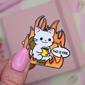 May include: A pin featuring a white cat illustration holding a yellow mug, surrounded by orange flames. The cat is in a burning situation, with the text bubble saying "This is fine." The pin has a black border.