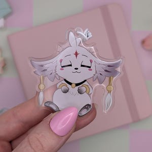 Könnte beinhalten: Ein klarer Acryl-Aufkleber mit einer weißen Cartoon-Figur mit geschlossenen Augen, Flügeln und goldenen Akzenten. Der Aufkleber hat einen schwarzen Kragen und ist vor einem rosa Hintergrund mit Blumen platziert.