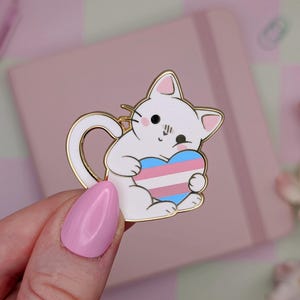 Op de afbeelding: Een witte kattenemaille pin met gouden rand, met een hart in de kleuren van de transgender pride vlag. De pin heeft een schattig ontwerp en staat op een roze achtergrond.