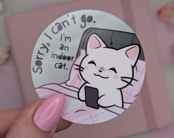 May include: A round sticker featuring a cartoon cat in bed, holding a phone. The cat is white with pink ears and a smiling face. Text on the sticker reads "Sorry, I can't go. I'm an indoor cat."