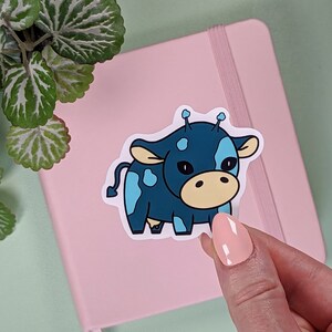 May include: A blue and yellow cartoon cow sticker with white spots. The cow has two antennas on its head and is looking to the right.