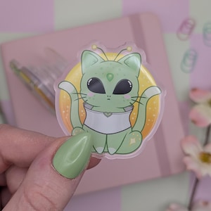 May include: A clear sticker of a green alien cat with large black eyes, antennae, and a white collar. The cat is set against a yellow and orange background. The sticker is held by a hand with a green painted nail.