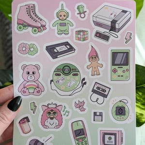 Op de afbeelding: Een stickerblad met diverse retro-thema illustraties. Ontwerpen omvatten een roze rolschaats, een groene Teletubby, een Rubik's Cube, een Nintendo console, een roze Care Bear, een Furby, een retro mobiele telefoon en een computer. De tekst "geekyfeels" staat onderaan.