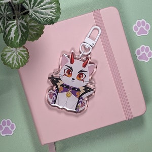 Peut inclure: Un porte-clés en acrylique transparent avec un chat blanc aux cornes rouges et un collier violet et or. Le porte-clés est suspendu à un carnet blanc avec une couverture rose.