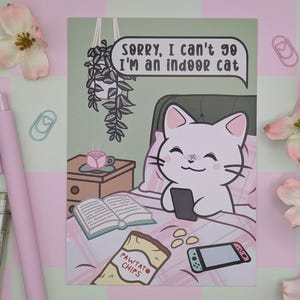 May include: A whimsical illustration of a white cat in bed, holding a phone. The text reads, "Sorry, I can't go I'm an indoor cat." A bag of "Pawtato Chips" and a game console are also visible. Pink flowers and pens surround the image.