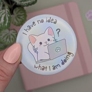 Op de afbeelding: Een blauwe en witte, iriserende knop met een witte kat die aan een laptop zit. De kat heeft een verwarde uitdrukking en de knop zegt "Ik heb geen idee wat ik doe?"
