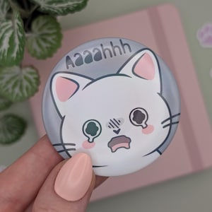Könnte beinhalten: Ein runder, grauer Knopf mit einer weißen Katze mit großen Augen und einem überraschten Ausdruck. Der Knopf trägt den Text "Aaaaahhh".