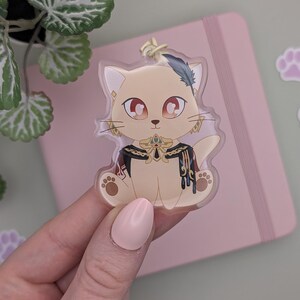 May include: A beige cat-shaped acrylic charm with red eyes, gold accents, and a feather. The charm is held in a hand, with a pink notebook and paw print stickers in the background. The cat charm has a decorative collar.