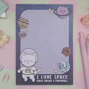 Puede incluir: Bloc de notas con temática de gatos espaciales. El bloc de notas tiene un fondo morado claro con ilustraciones de gatos con trajes espaciales y planetas. El texto "I LOVE SPACE (both outer & personal)" está en la parte inferior. Un bolígrafo rosa y flores están al lado.