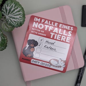 May include: A red and white emergency contact card for pets, with illustrations of a dog and cat. The card, with the text "IM FALLE EINES NOTFALLS TIERE", rests on a pink notebook, with a black pen and marker nearby.