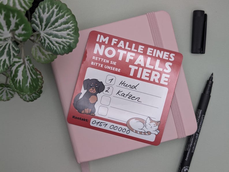 May include: A red and white emergency contact card for pets, with illustrations of a dog and cat. The card, with the text "IM FALLE EINES NOTFALLS TIERE", rests on a pink notebook, with a black pen and marker nearby.