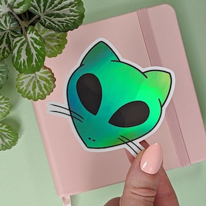 Op de afbeelding: Een holografische sticker van een groene alienkat met grote zwarte ogen. De sticker zit op een roze notitieboek.