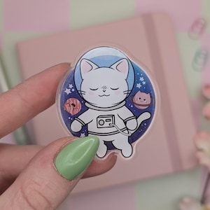 May include: A clear, round acrylic pin depicting a white cat astronaut in space. The cat is wearing a spacesuit and helmet, with planets and stars in the background. The pin is held by a person with green nail varnish.