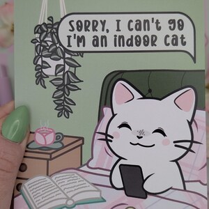 May include: A greeting card with a cartoon cat in bed holding a phone. The cat is white with pink ears and cheeks. Text in a speech bubble reads, "Sorry, I can't go I'm an indoor cat." A small table has a cup of tea and a book.