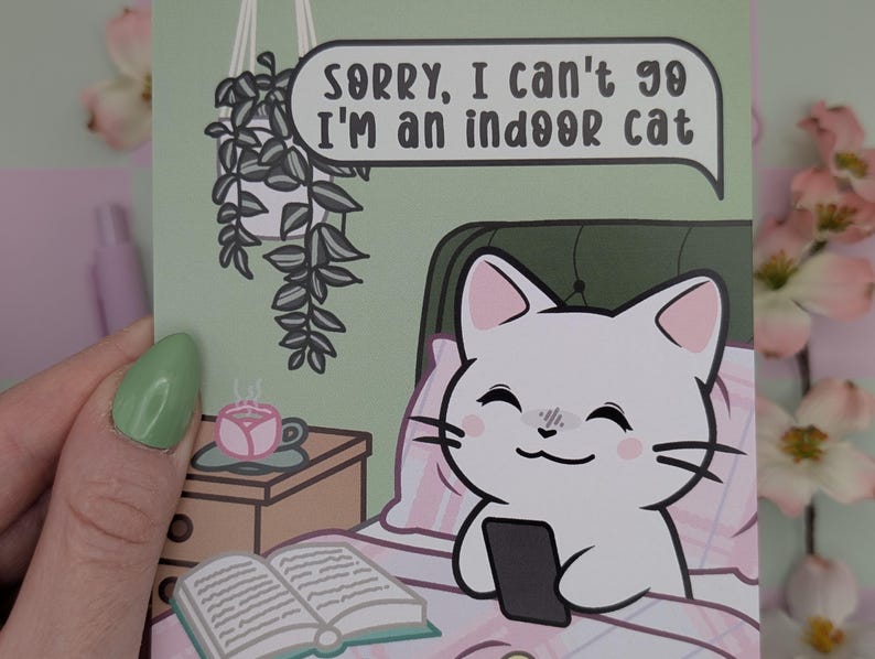 May include: A greeting card with a cartoon cat in bed holding a phone. The cat is white with pink ears and cheeks. Text in a speech bubble reads, "Sorry, I can't go I'm an indoor cat." A small table has a cup of tea and a book.