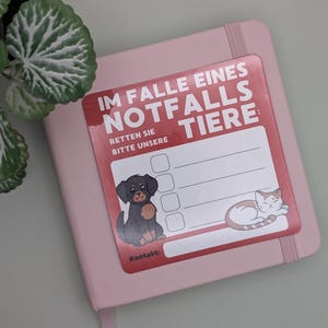 May include: A pink notebook with a sticker that reads "IM FALLE EINES NOTFALLS TIERE: RETTEN SIE BITTE UNSERE" in red and white. The sticker features a cartoon dog and cat. A green plant is in the upper left corner.