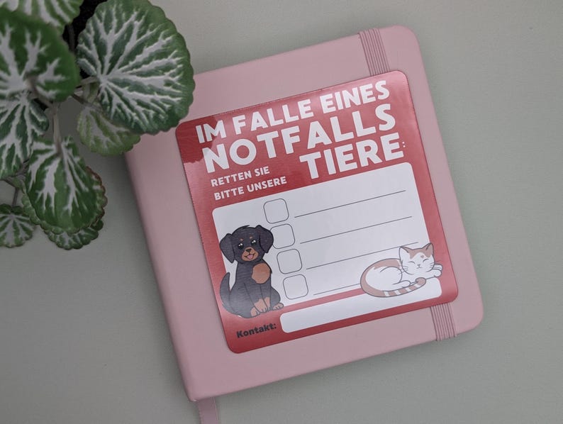 May include: A pink notebook with a sticker that reads "IM FALLE EINES NOTFALLS TIERE: RETTEN SIE BITTE UNSERE" in red and white. The sticker features a cartoon dog and cat. A green plant is in the upper left corner.