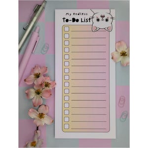 Puede incluir: Un bloc de notas rectangular con un fondo degradado rosa y amarillo, con el texto "My endless To-Do List" y una ilustración de gato de dibujos animados. Bolígrafos rosas, clips y flores están dispuestos alrededor del bloc de notas.